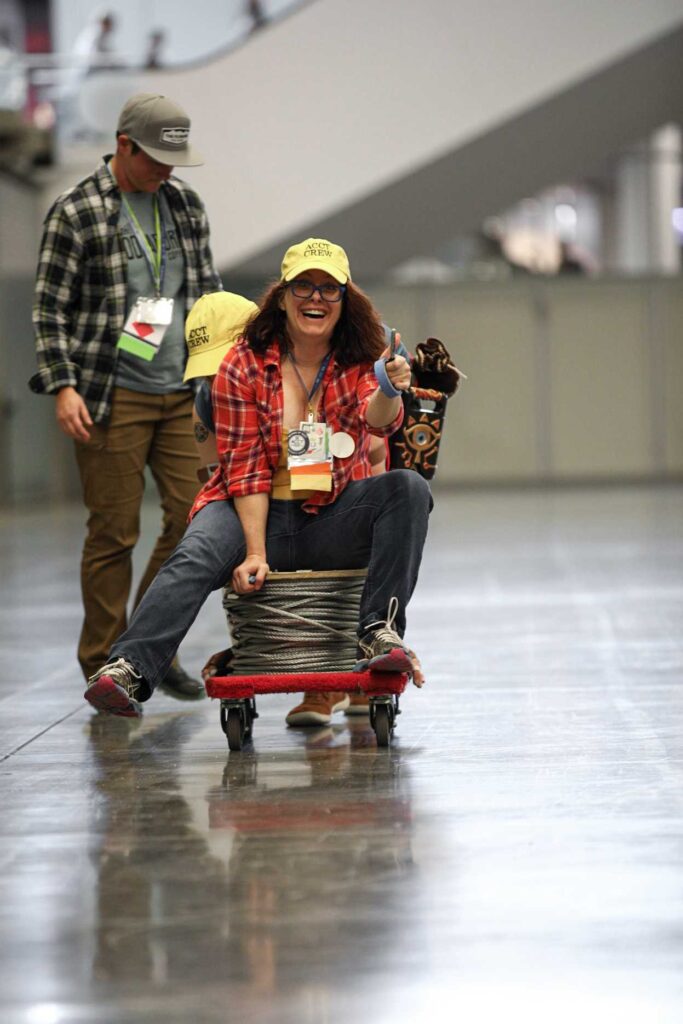 A member of the Service Crew smiles and holds a thumbs up while riding a small cart that transports the galvanized steel aircraft cable from one side of the Exhibit Hall to the other during the Harnessing ACCT showdown hosted at the 2025 conference and exposition.