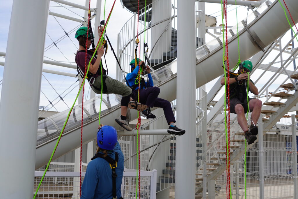 Three people wearing helmets and full body harnesses practice their rope access skills at a local challenge course under the supervision of one of the session presenters, who is watching from the ground while helping manage ropes.