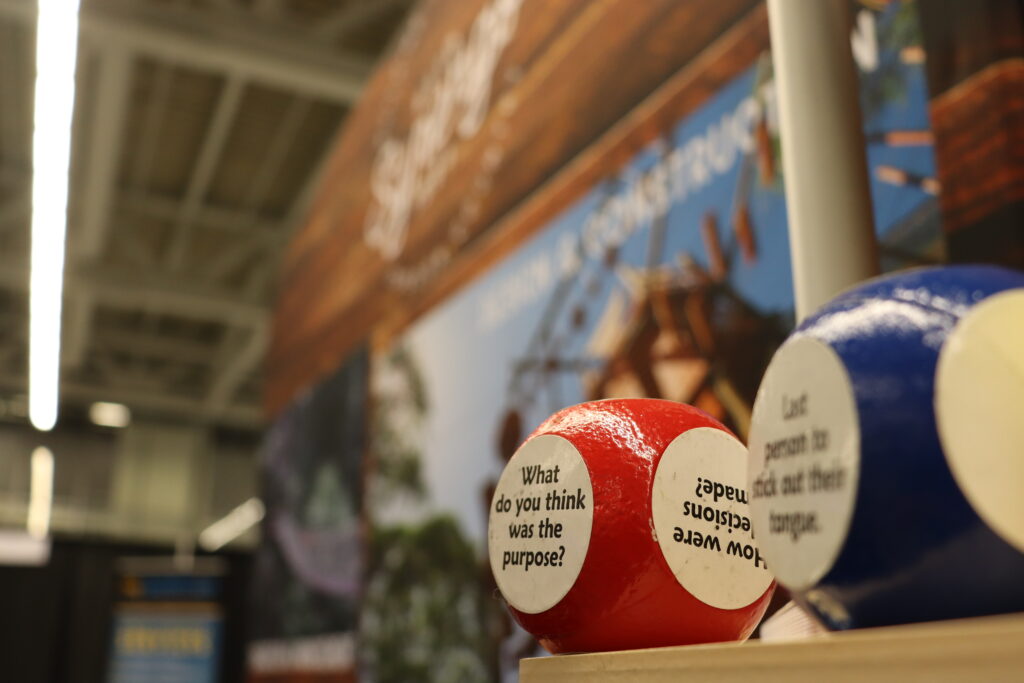 A question debrief ball that currently displays the question, "What do you think was the purpose?" is featured while an exhibitor booth is blurred in the background.