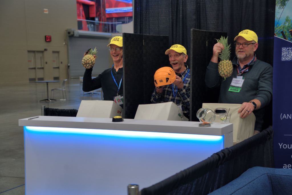 Three volunteers from the Service Crew smile as they hold up a pineapple, a helmet, and another pineapple during the human slot machine at the ACCT Booth in the Exhibit Hall at the 2025 conference and exposition.