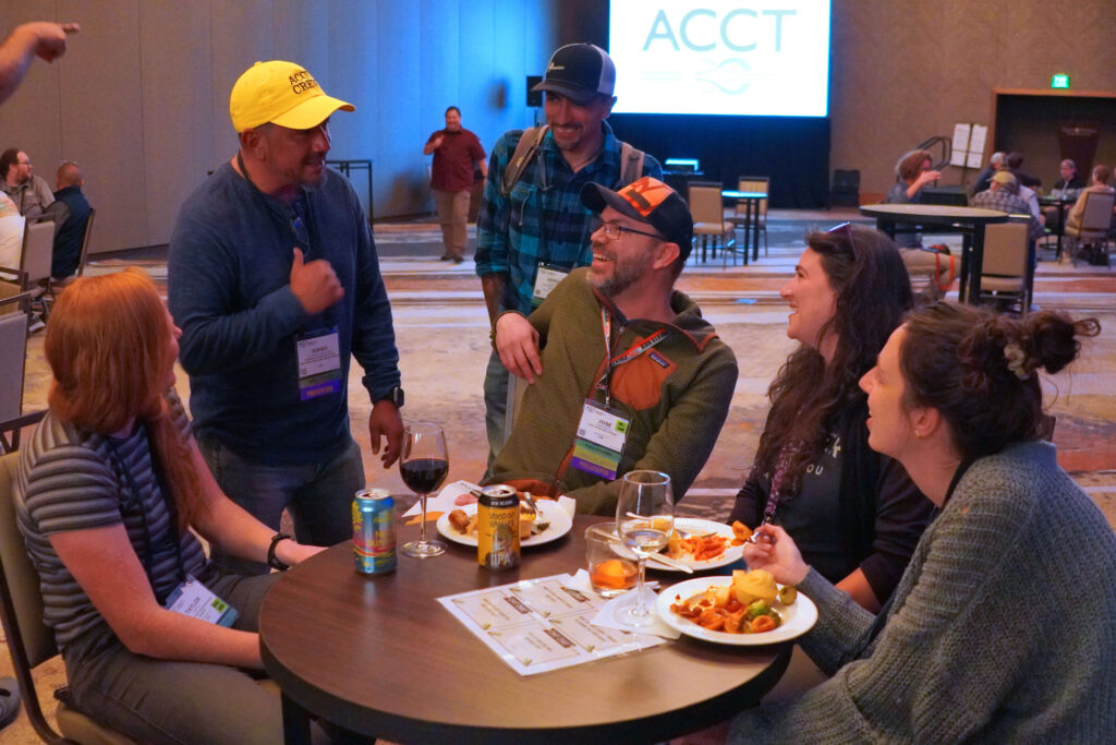 Six people, mostly sitting around a small round table filled with food and drinks, laugh and smile with a Service Crew member wearing their yellow hat as the ACCT brand logo is seen on a projector screen in the background.