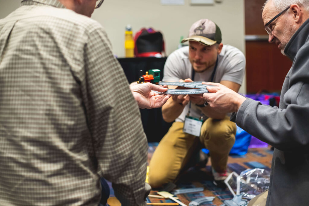 Four people work together to balance a toy train on its track as they hold it in the air while attempting to get the train all the way around the track without falling during a workshop.