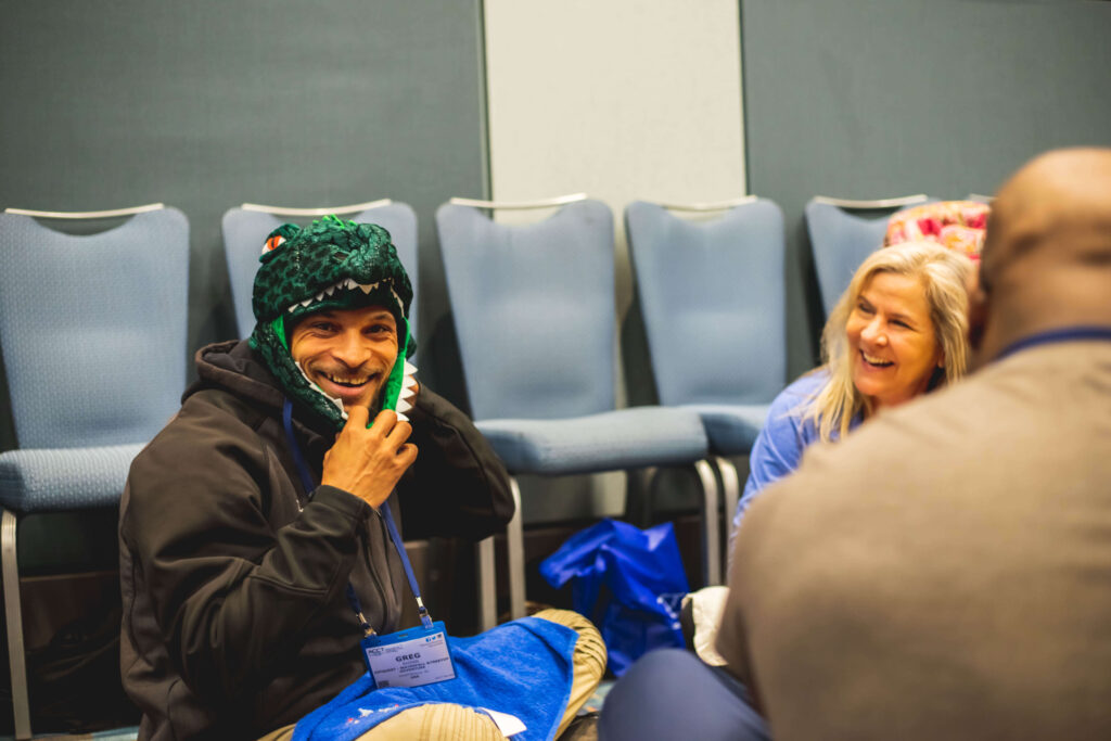 A woman smiles at a man wearing a dinosaur hat as he smiles at the camera, which is looking over the shoulder of another man in the group as they sit on the floor of a workshop.