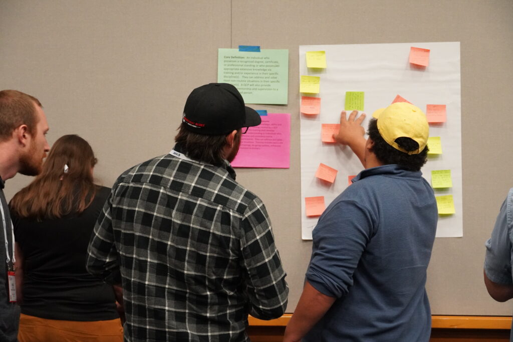 Three attendees review what people have written on sticky notes, which is hanging on a giant sheet of butcher paper taped to the wall of a workshop as other attendees are seen milling around.