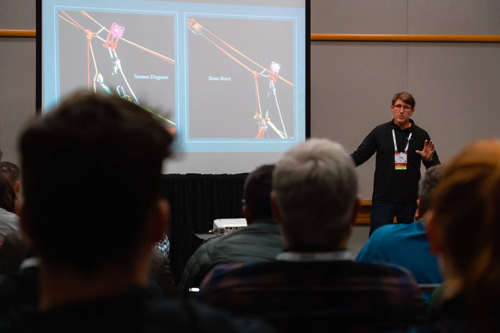 A room full of attendees look at two images seen on a slideshow presentation while the speaker discusses their content.