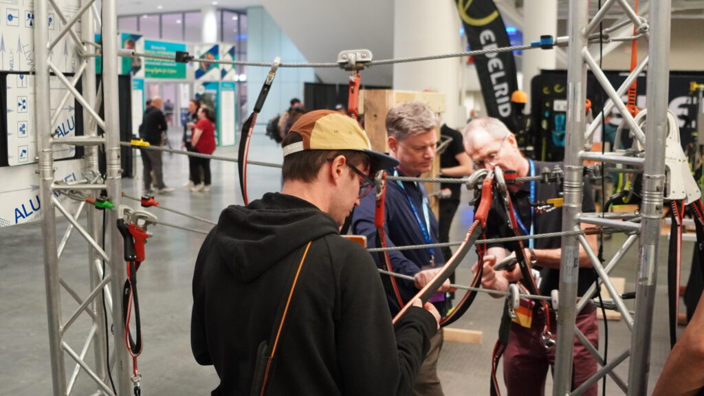 Two people examine the CLiC-iT belay system at their booth in the Exhibit Hall during the 2025 conference and exposition. One person is being shown the equipment by CLiC-iT staff.