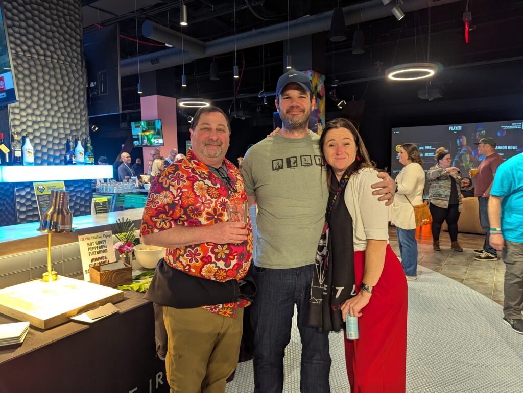 Three people smile as they hug one another near one of the food stations available at the After Hours Social sponsored by Granite Insurance during the 2025 ACCT conference and exposition.