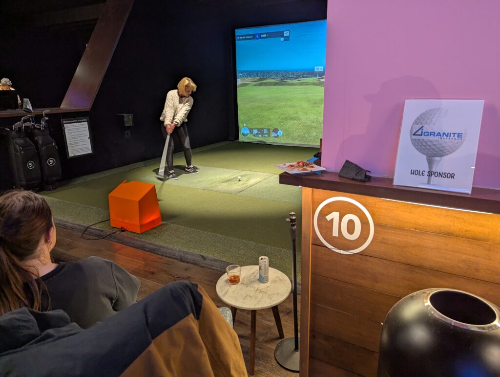 A woman sits in a cushioned chair as she watches another woman use a golf simulator at the After Hours Social sponsored by Granite Insurance during the 2025 ACCT conference and exposition.