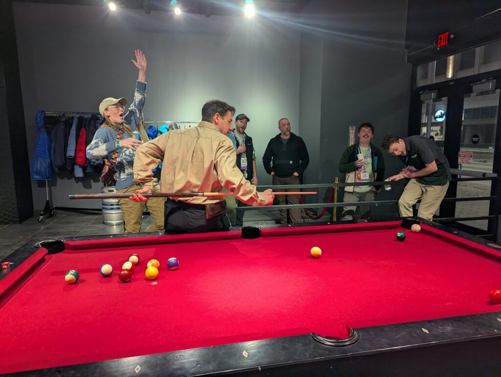 A crowd of people react to an attendee successfully sinking a pool ball while holding the cue stick behind his back at Five Iron Golf during the After Hours Social sponsored by Granite Insurance at the 2025 ACCT conference and exposition.