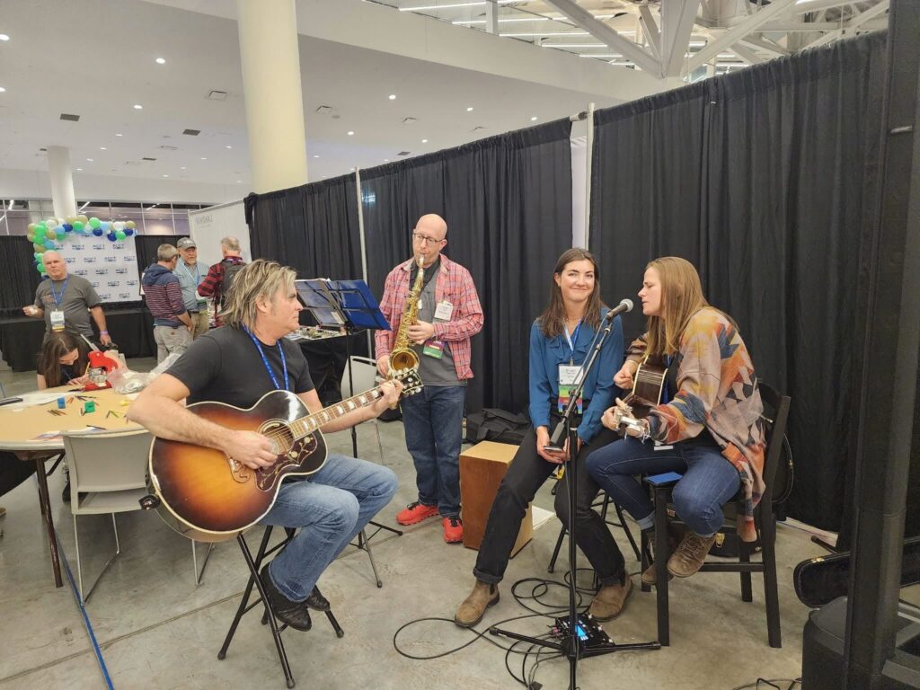 One man sits on a stool plays acoustic guitar while another stands and plays the saxophone. Next to them a woman sings and plays acoustic guitar while sitting in a chair as another singer sits next to her and smiles at the camera in the Exhibit Hall at the 2025 conference and exposition.