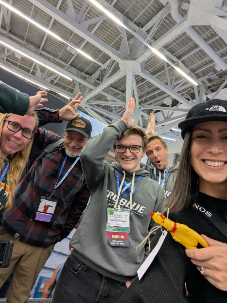 Three members of the Harnessing ACCT team Red Feather Flyers pretend to be chickens with two exhibitors from the Head Rush Technology booth in the Exhibit Hall while one person holds a small rubber chicken from the Association for Experiential Education booth during the 2025 conference and exposition.