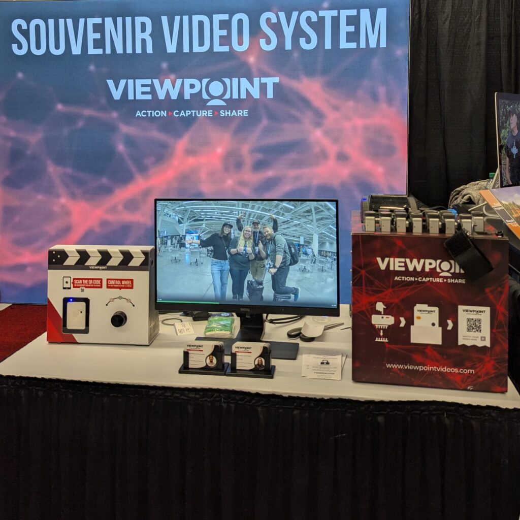 Four members of the Harnessing ACCT team Red Feather Flyers pose while the Viewpoint souvenir video system displays their image on a tv screen after collecting footage in the Exhibit Hall at the 2025 conference and exposition.