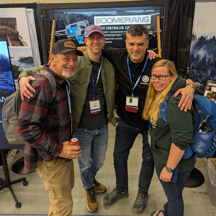 Two members of the Harnessing ACCT team Red Feather Flyers give a group hug to the two exhibitors of the Boomerang booth in the Exhibit Hall during the 2025 conference and exposition.