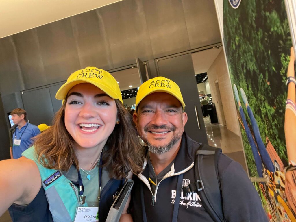 Two people wearing the iconic yellow Service Crew hats smile at the camera outside of the Exhibit Hall.