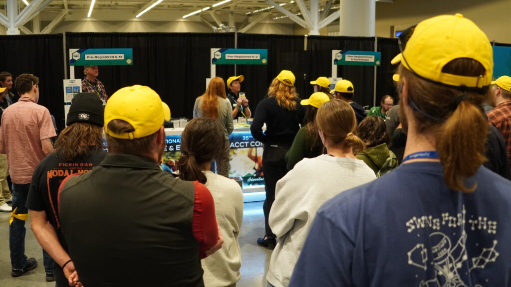 A crowd of Service Crew volunteers wearing the iconic yellow hat listens to Service Crew Coordinator Leigh Carruth explain how badge pick-up at registration works during Service Crew Orientation at the 2025 ACCT conference and exposition.