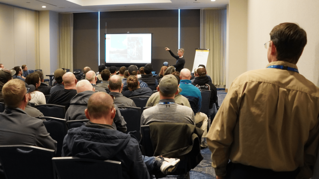 A presenter explains a series of photos seen on a projector screen to a crowd of session attendees, most of whom are sitting but a few folks stand along the sidelines at the 2025 ACCT conference and exposition.