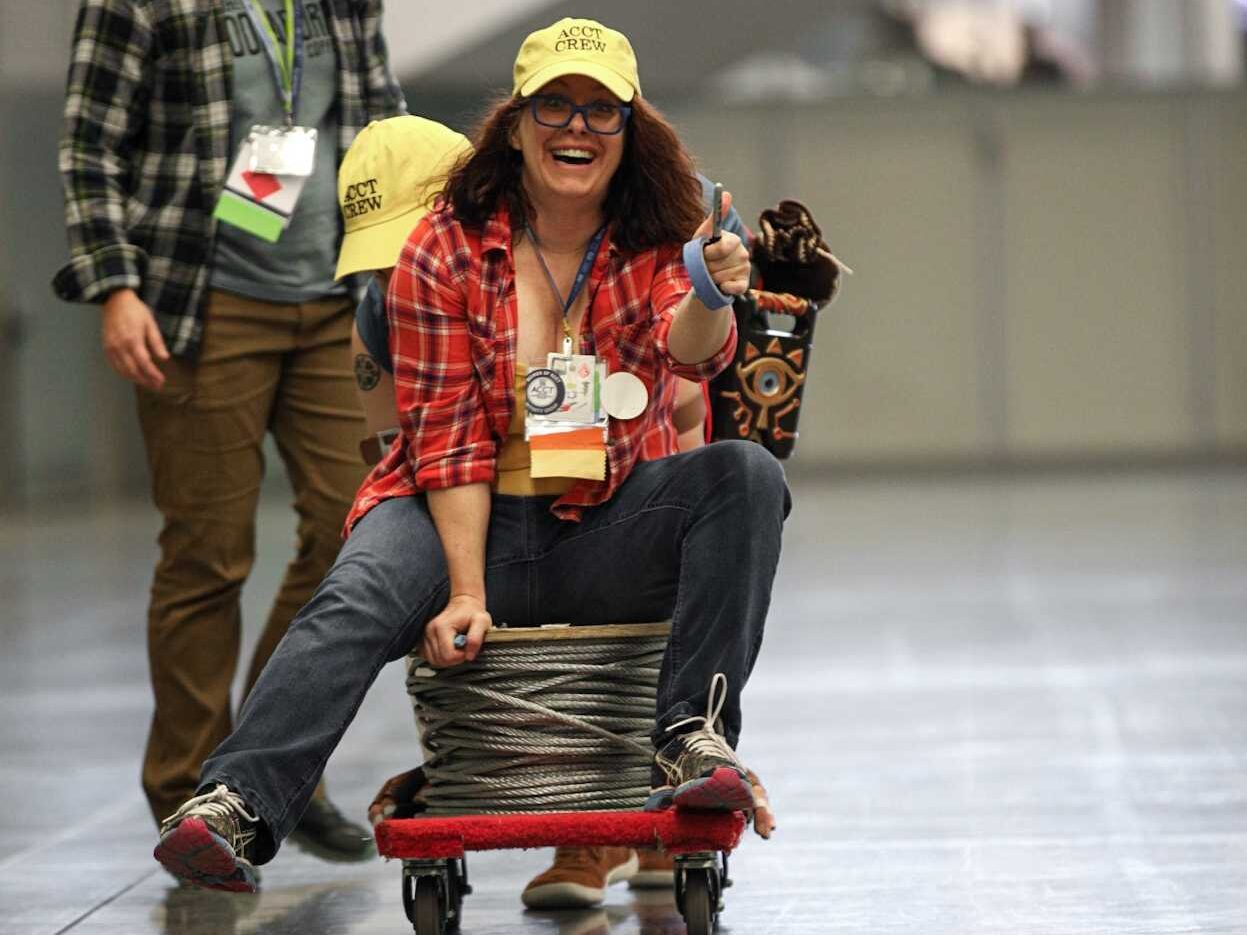 A member of the Service Crew smiles and holds a thumbs up while riding a small cart that transports the galvanized steel aircraft cable from one side of the Exhibit Hall to the other during the Harnessing ACCT showdown hosted at the 2025 conference and exposition.