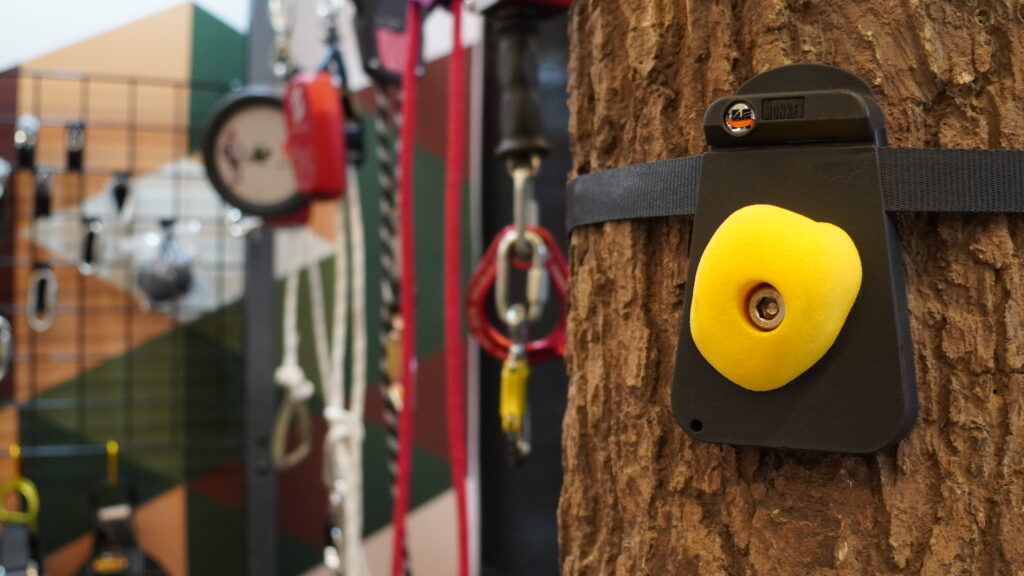 The photo focuses on a modular rock hold strapped to a tree while various pieces of equipment are seen in the background, taken in the Exhibit Hall at the 2025 ACCT conference and exposition.